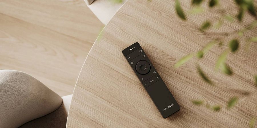 T&eacute;l&eacute;commande avec &eacute;cran OLED du syst&egrave;me home cin&eacute;ma sans fil Loewe We. BOOST, pos&eacute;e sur une table en bois clair.